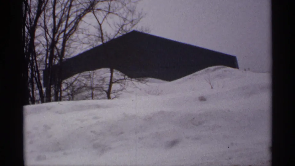 1971 Wisconsin 8mm frame showing a dark outbuilding or roofline behind deep snowdrifts, with bare trees in the winter background.
