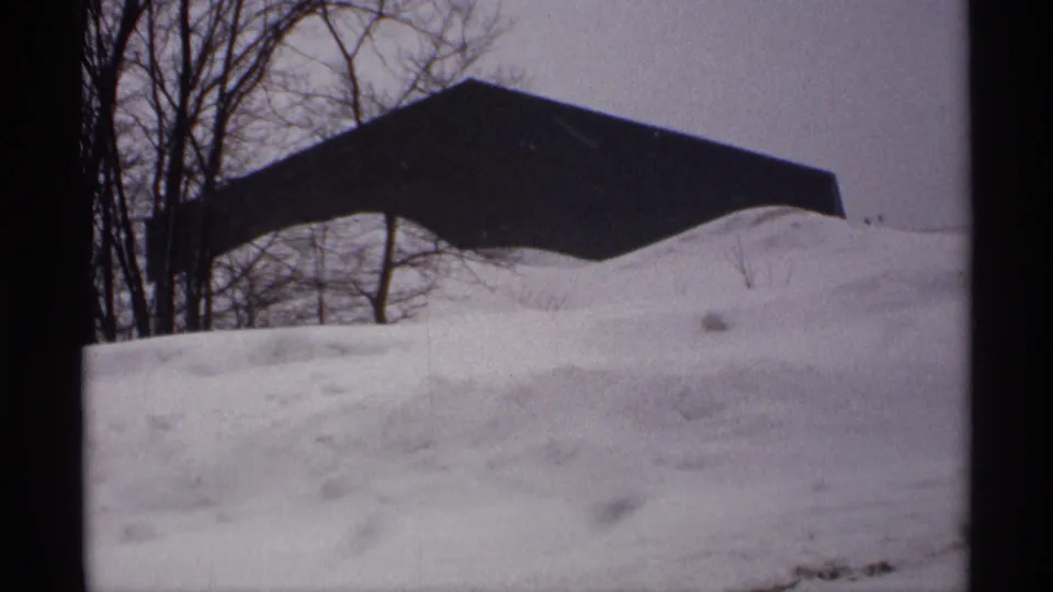1971 Wisconsin winter frame showing a dark angular outbuilding or roof form partly buried by snow, with leafless trees beyond.