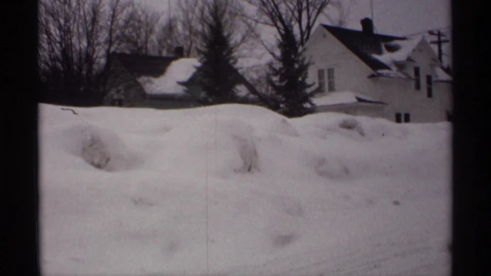 1971 Wisconsin 8mm frame with snow-covered residential houses, bare trees, and a tall snowbank across the foreground.