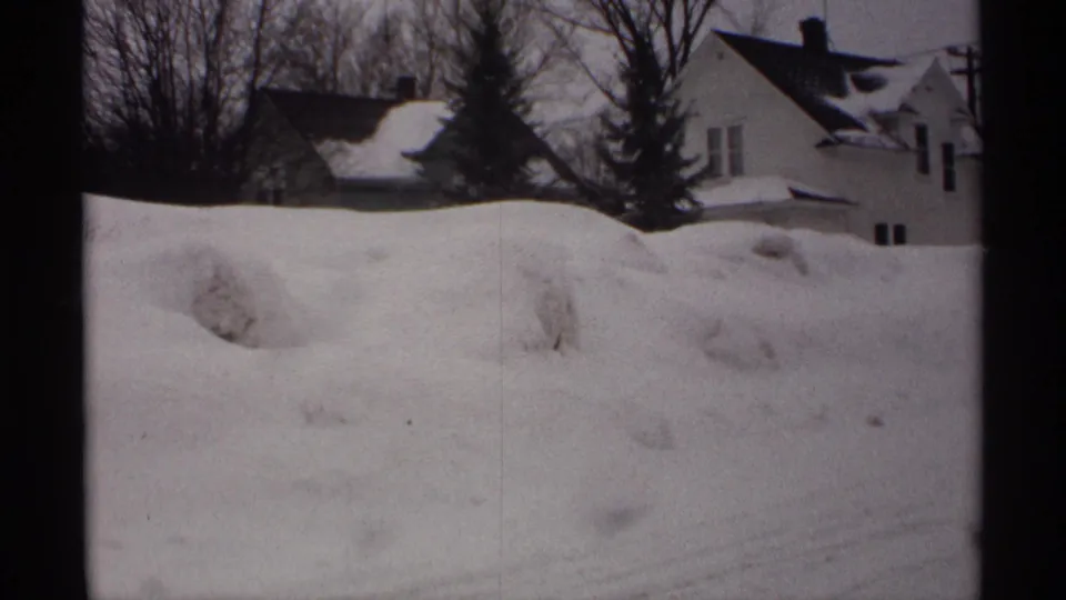 1971 Wisconsin home-movie frame showing a white house, roof snow, bare trees, and deep snow piled along the street or driveway edge.