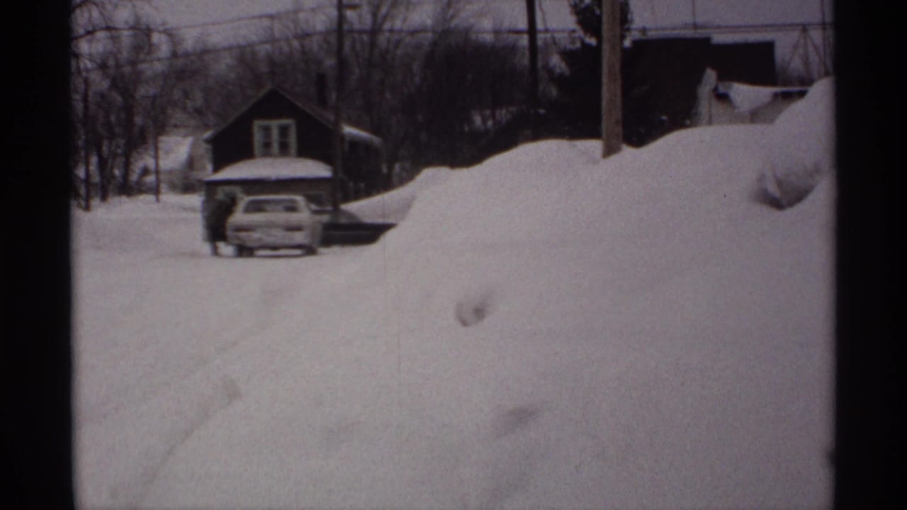 Snow-covered residential street with a partially buried car and a person standing nearby.