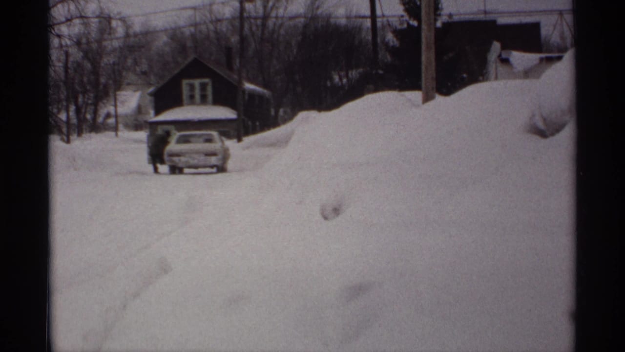 1971 Wisconsin home-movie view of high snowbanks along a residential street, with a car, house, bare trees, and utility lines visible.