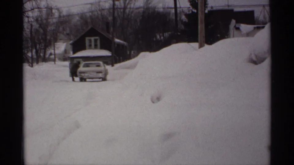 1971 Wisconsin home-movie view of high snowbanks along a residential street, with a car, house, bare trees, and utility lines visible.