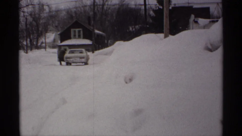 1971 Wisconsin 8mm frame showing a person near a partially snow-buried car, with a small house and heavy snowdrifts around the street.