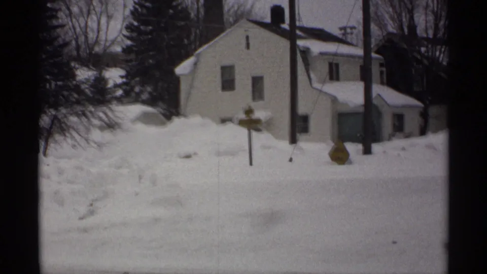 1971 Wisconsin 8mm frame of a white house, deep snowbanks, utility poles, bare trees, and a yellow road sign on a winter street.