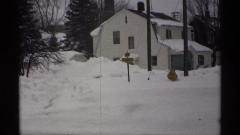 1971 Wisconsin 8mm frame showing a white house, roadside signs, poles, bare trees, and deep snowbanks.