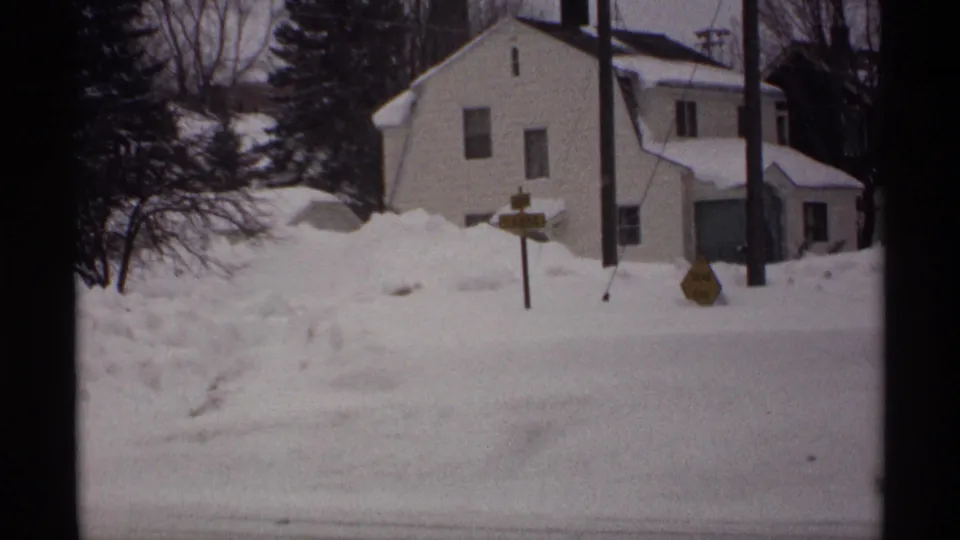 1971 Wisconsin winter view of a white house, yellow road signs, utility poles, and piled snow beside the street.