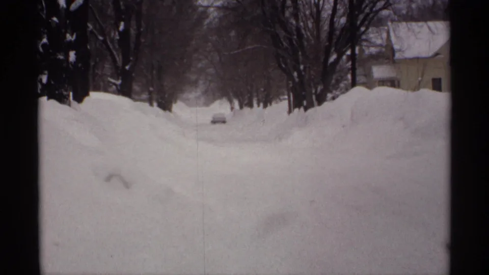 1971 Wisconsin winter road view with high snowbanks, bare trees, nearby houses, and a small distant car.