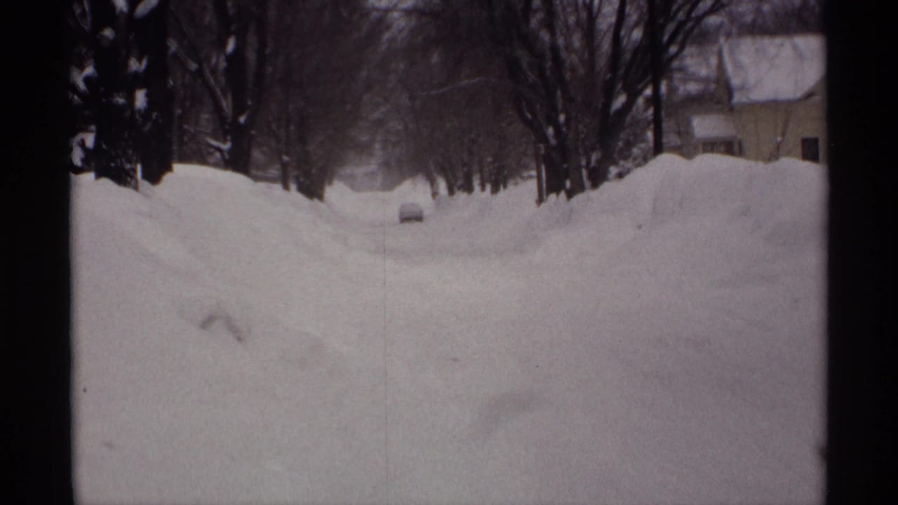 Snow-packed residential street with high snowbanks, bare trees, and a distant car.