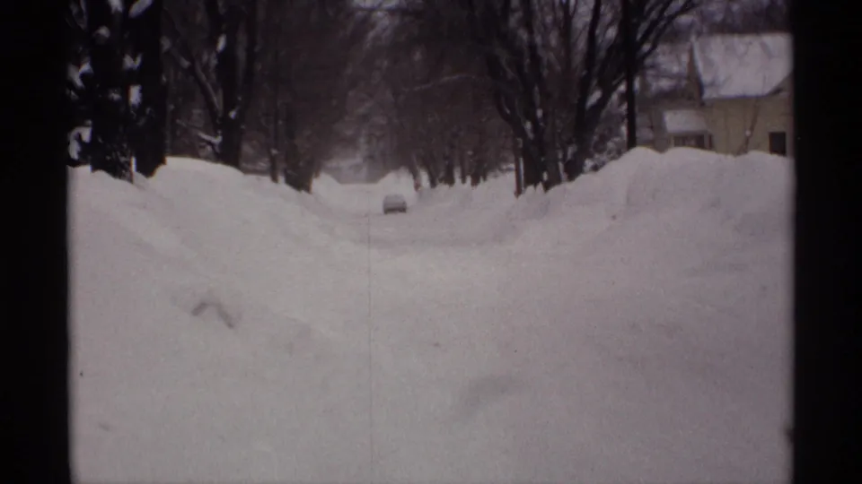 1971 Wisconsin 8mm frame of a snow-covered residential street lined with high snowbanks, leafless trees, and a distant car.