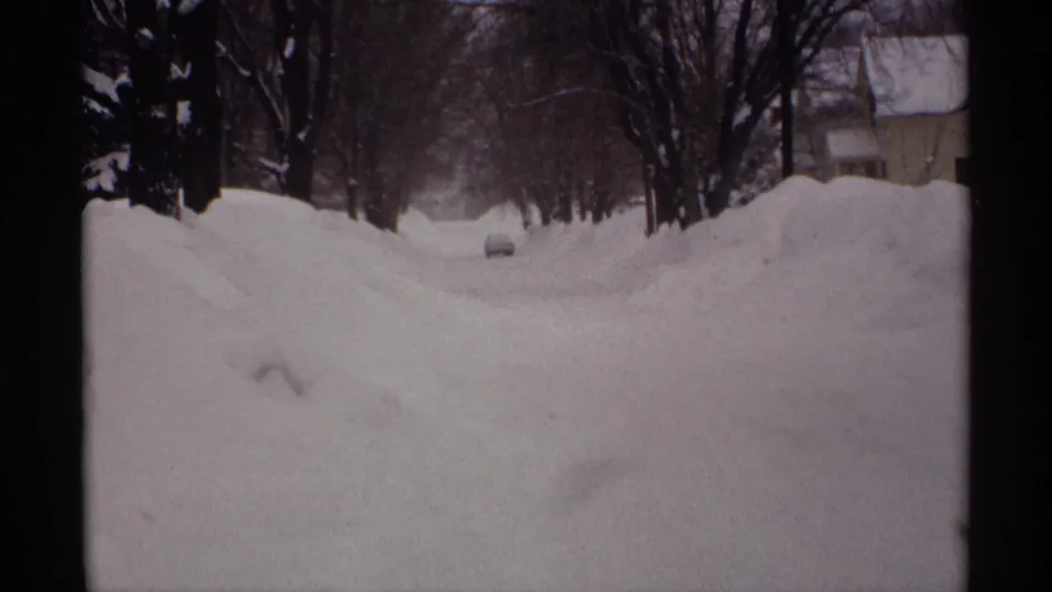 1971 Wisconsin home-movie view down a snowbound residential street, with a car ahead, bare trees, and houses partly visible through the snow.