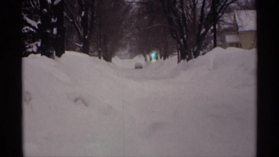 1971 Wisconsin 8mm frame showing a snow-covered residential road with high banks, bare trees, and faint headlights in the distance.
