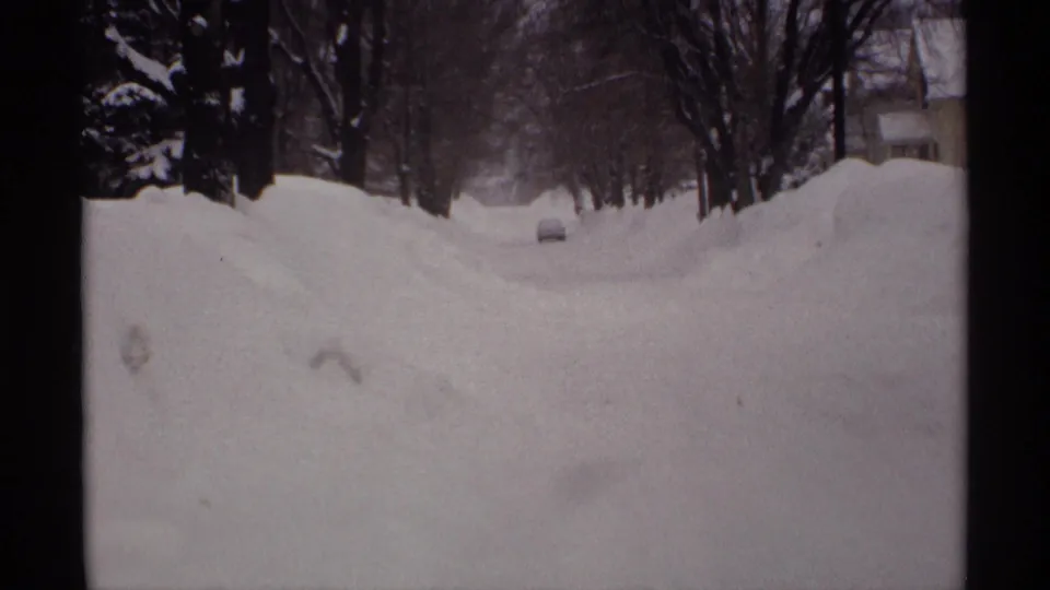 1971 Wisconsin frame of a quiet residential street buried in snow, with a car ahead, tall snowbanks, and leafless trees on both sides.