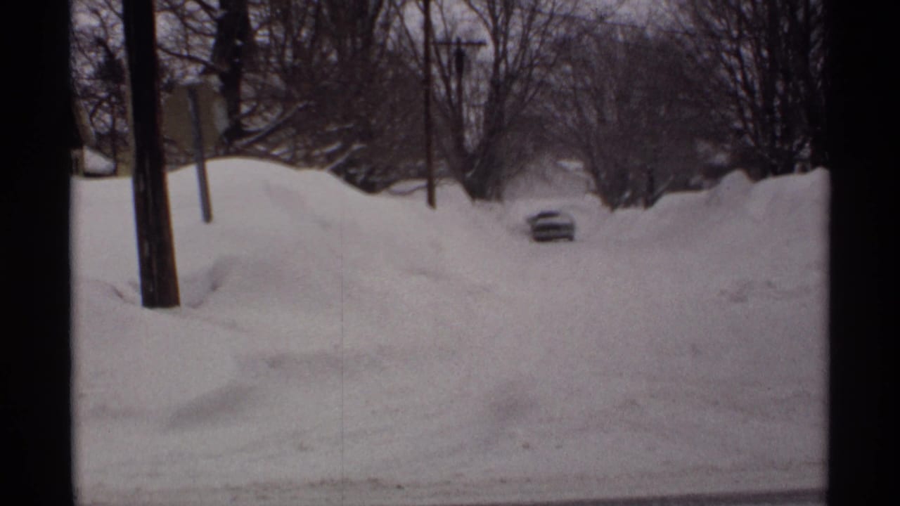 Deep snowbanks along a residential street with a partly buried car in the distance.