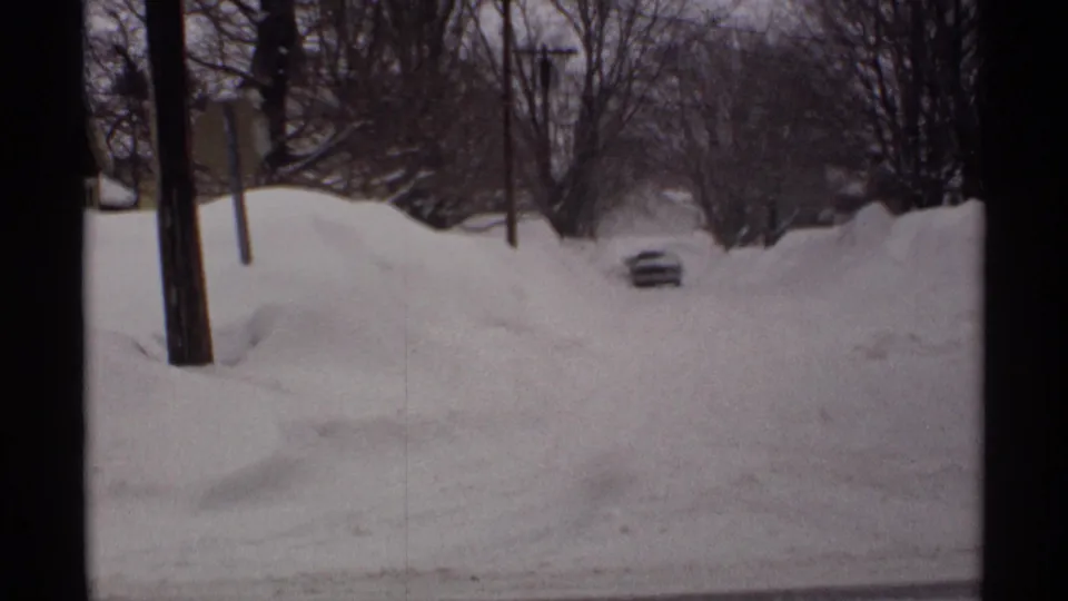 1971 Wisconsin 8mm frame showing deep snowbanks, bare trees, utility poles, and a car partly buried at the end of a residential street.