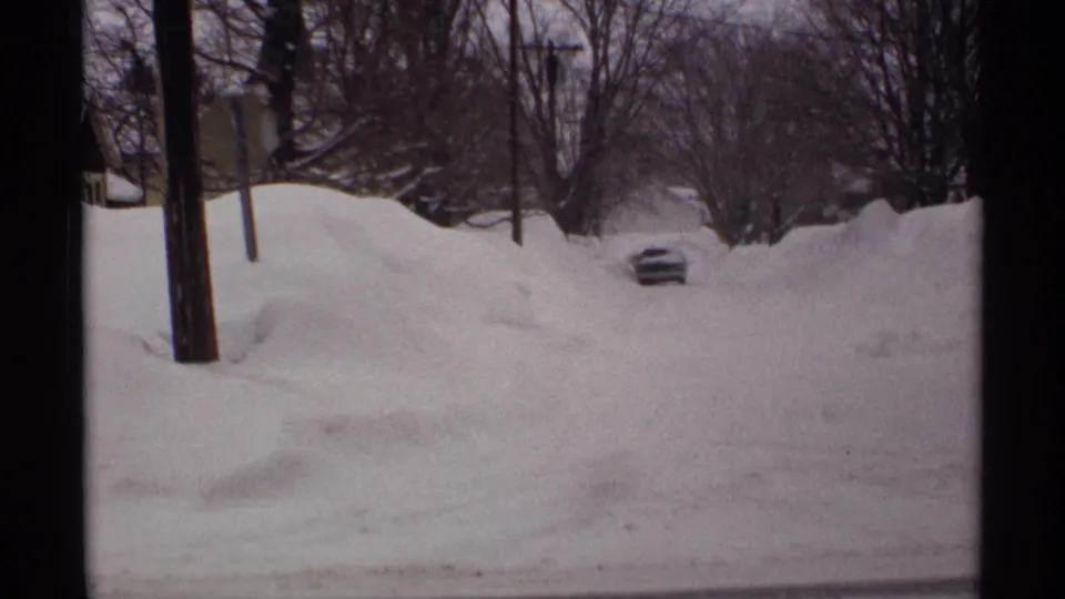 A 1971 Wisconsin winter street appears nearly blocked by high snowdrifts, with a car partly visible beyond bare trees and utility poles.