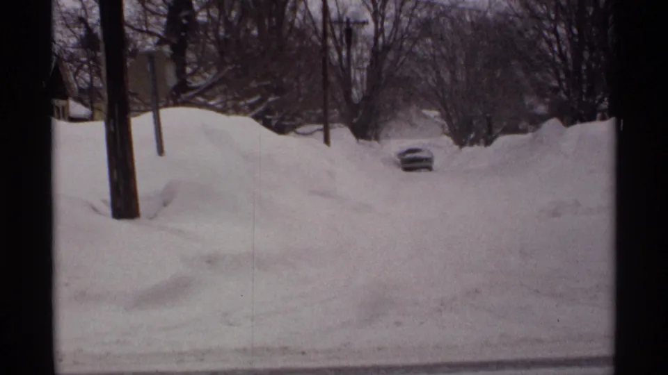 1971 Wisconsin home-movie frame of a quiet residential street overwhelmed by deep snow, with a car still visible above the drifts.