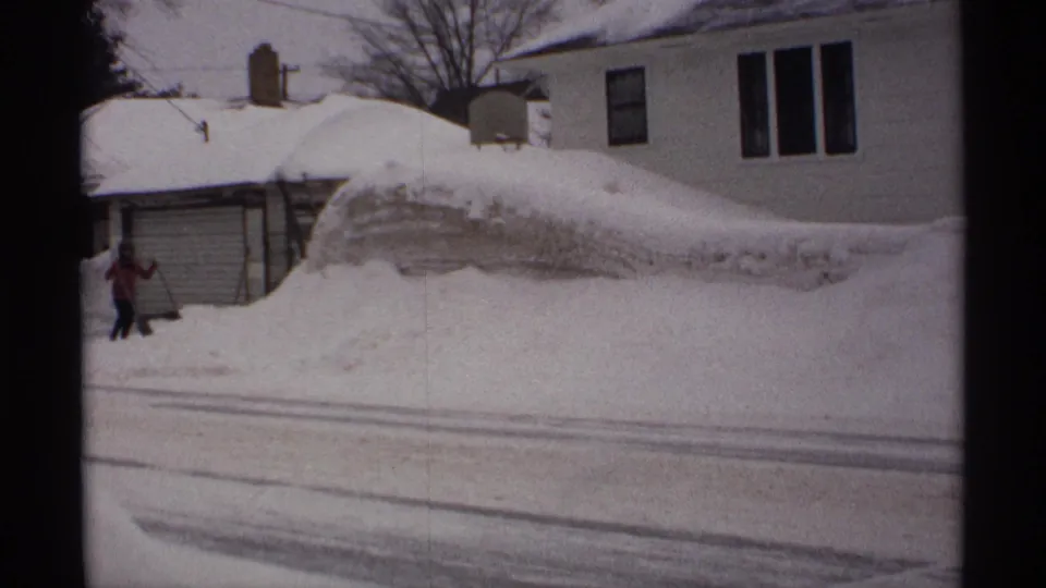 In a 1971 Wisconsin residential scene, a person in a red jacket shovels near a garage while tall snowbanks line the street.