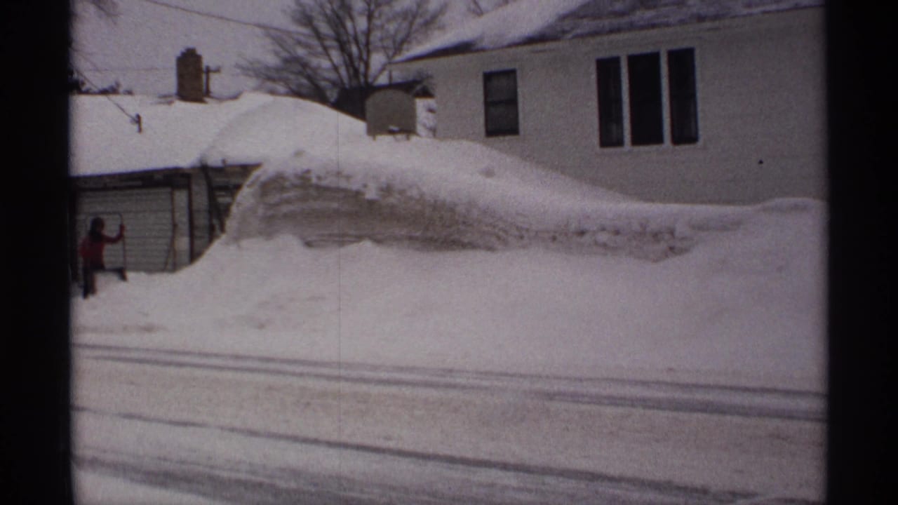 Snowbank outside a house with a person in a red jacket near a garage.