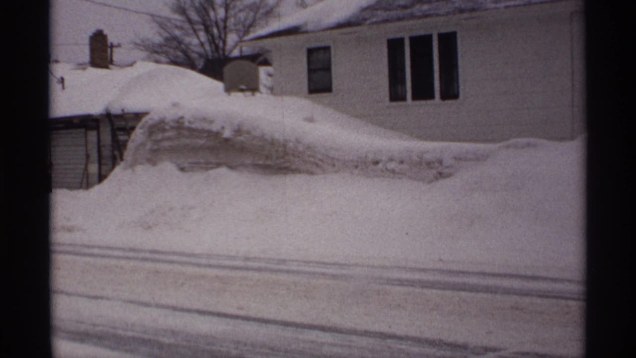 Large snow mound in front of a house and garage on a snowy street.