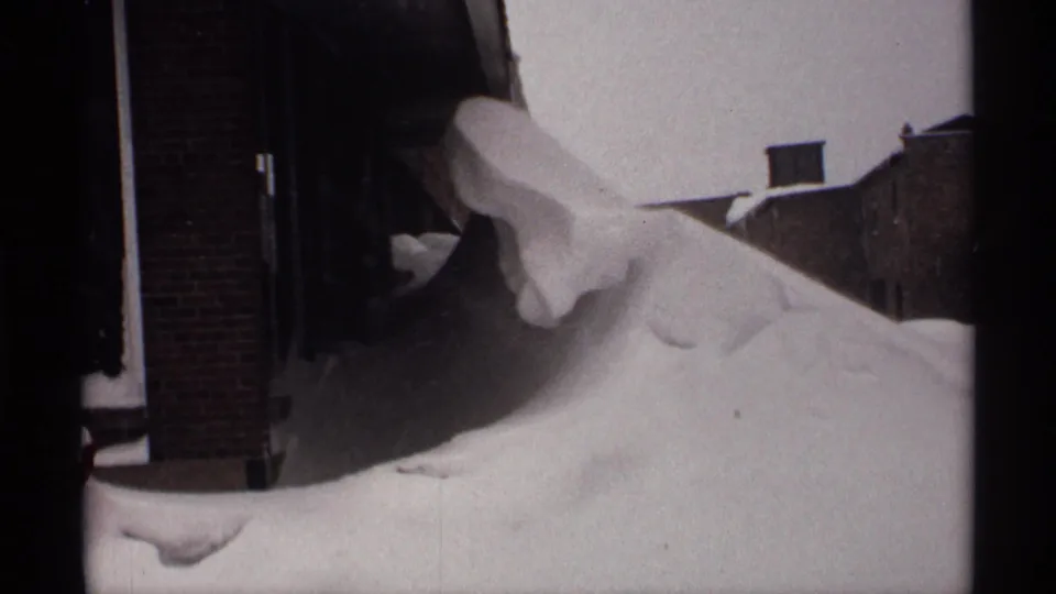 A close residential view shows a heavy snowdrift pressed against a brick wall and doorway in vintage 8mm winter footage.
