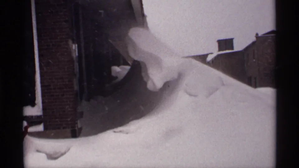 1971 Wisconsin 8mm frame showing snow heaped near brick residential buildings, with dark film borders and a narrow street view.