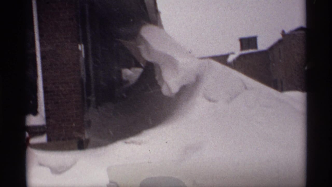 Large snowbank pressed against brick houses with rooftops visible beyond.