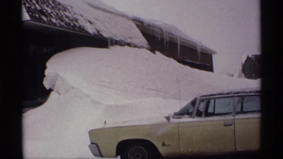 1971 Wisconsin 8mm still showing icicles on a snowy roof and a light-colored station wagon partly buried by a large drift.