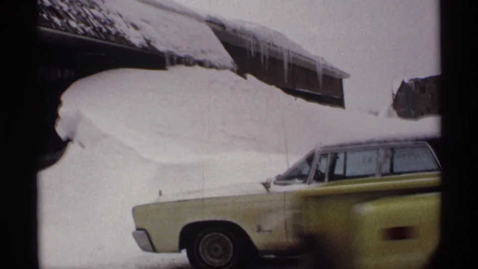 1971 Wisconsin home-movie frame of a station wagon parked beside a deep snowbank, with icicles hanging from the nearby roof.