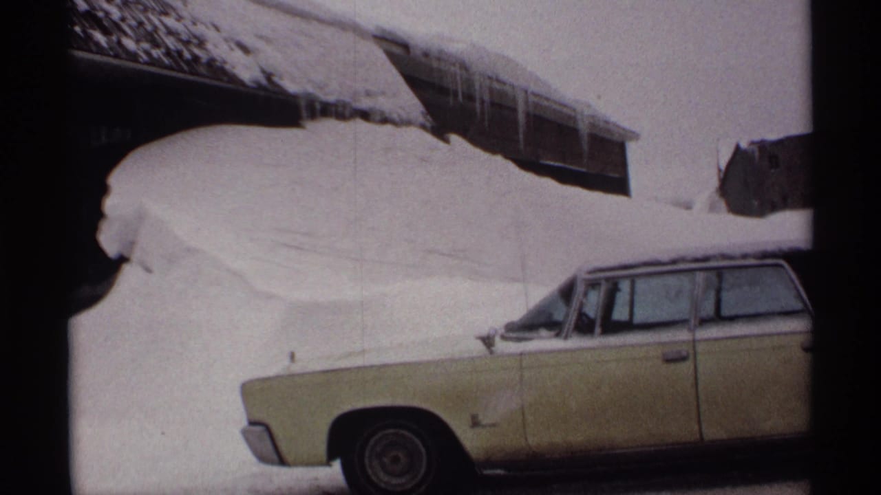 Snow-buried pale yellow station wagon beside a building with heavy roof snow.