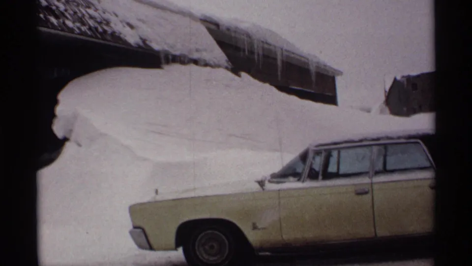 1971 Wisconsin 8mm frame showing a pale yellow station wagon nearly surrounded by deep snow below snow-loaded eaves.