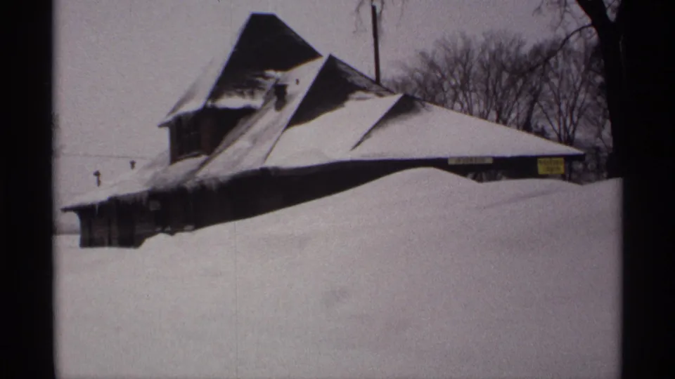 1971 Wisconsin 8mm frame of a steep-roofed building nearly covered by deep snow, with bare trees and a small sign visible.