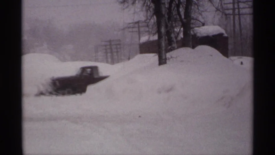 1971 Wisconsin 8mm frame with a car partly buried or blurred by motion near high snowbanks, trees, and utility poles.