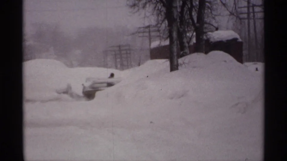 1971 Wisconsin home-movie frame showing a person working near a buried car amid high snowbanks, trees, and utility poles.