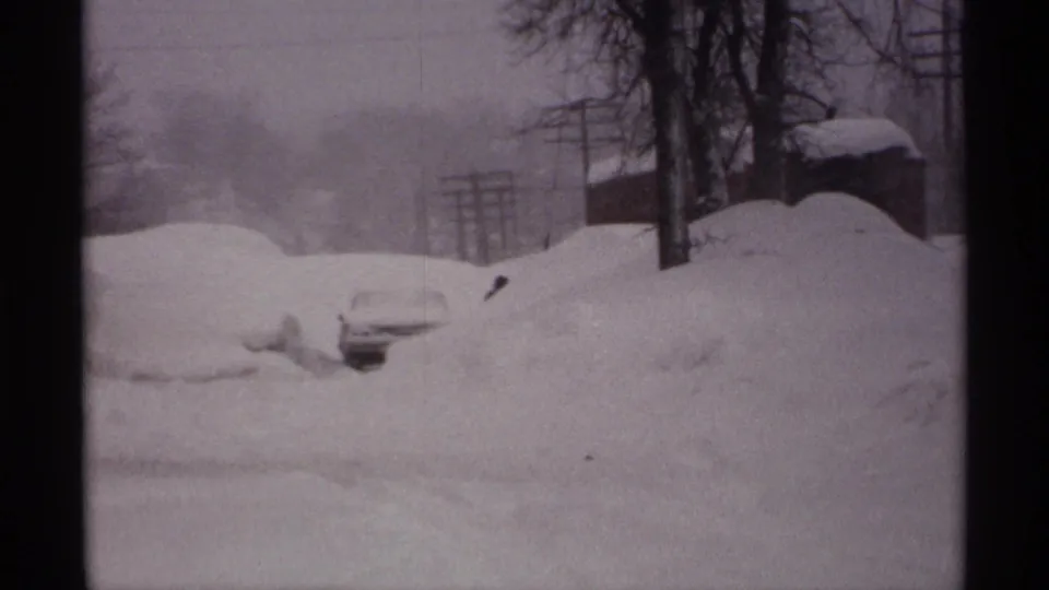 1971 Wisconsin 8mm frame showing a snowbound residential street with partly buried cars, utility poles, bare trees, and distant houses.