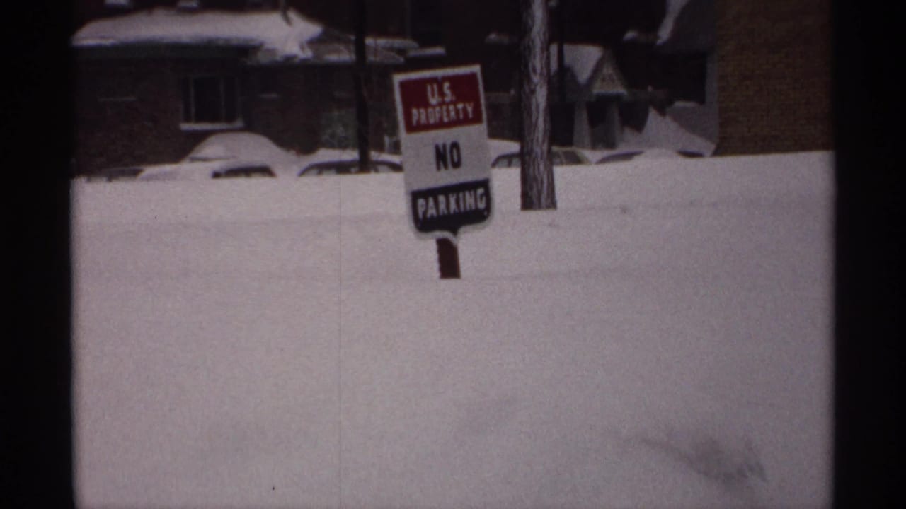 U.S. Property No Parking sign stands in deep snow near houses and a buried car.