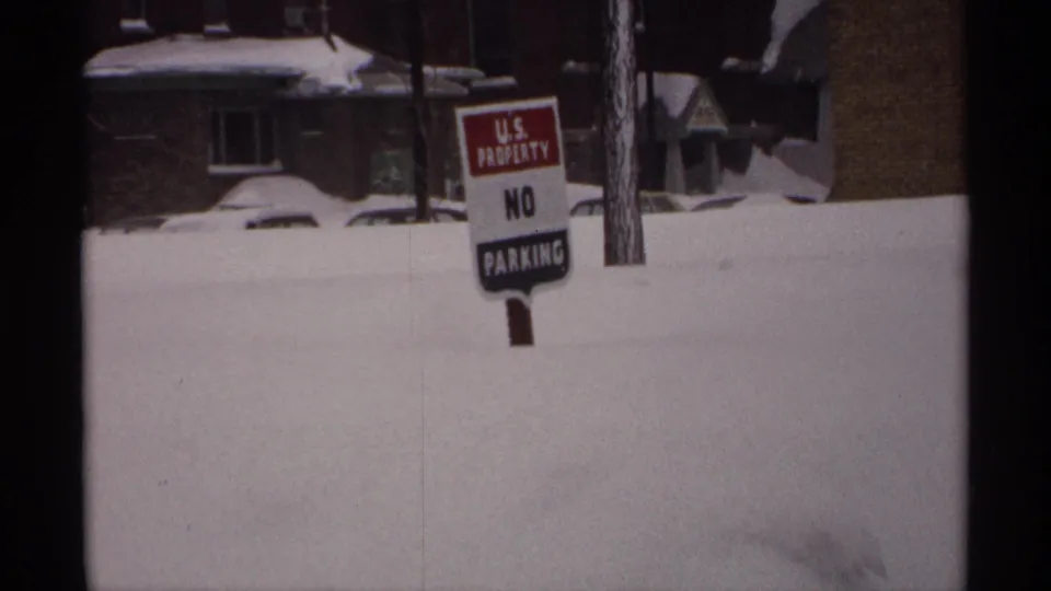 1971 Wisconsin 8mm frame showing a No Parking sign in deep snow, with houses, a tree trunk, and a buried car behind it.