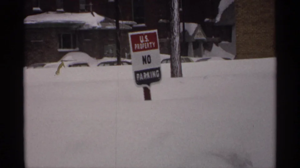 1971 Wisconsin 8mm frame of a U.S. Property No Parking sign in a deep snowbank, with houses and a buried car behind it.