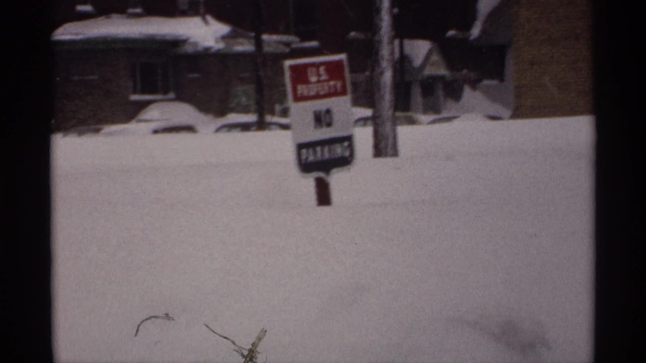 Snow-covered roadside with a U.S. property no parking sign and houses behind deep drifts.