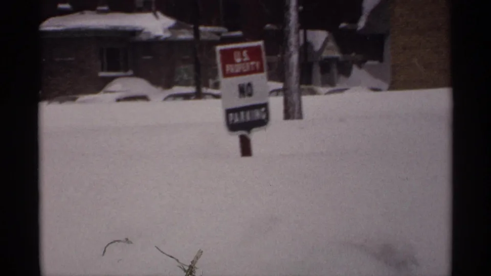 1971 Wisconsin 8mm frame showing a snowbound roadside, a U.S. property no parking sign, brick homes, and deep winter accumulation.