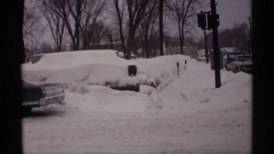 1971 Wisconsin street scene with parked cars partly buried by snow, high plowed banks, traffic lights, and leafless winter trees.