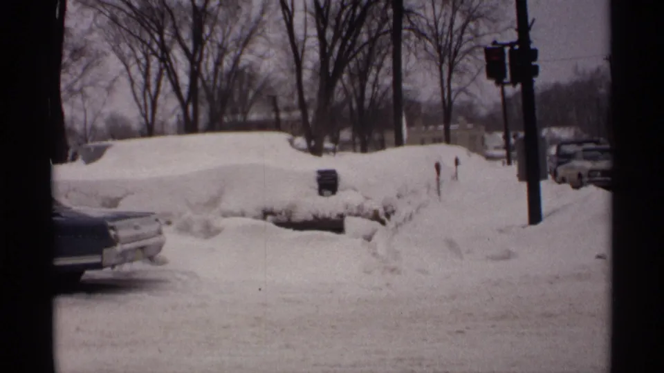 1971 Wisconsin 8mm view of a snowy street corner with buried cars, tall roadside snowbanks, a traffic signal, and bare trees.