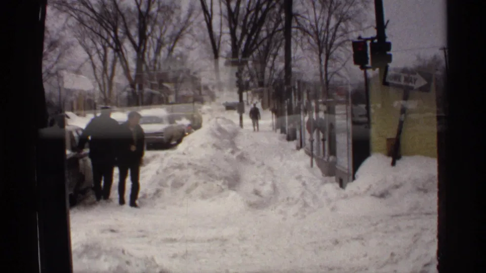 1971 Wisconsin town sidewalk with pedestrians walking beside snowbanks, parked cars, storefronts, and a visible one way street sign.