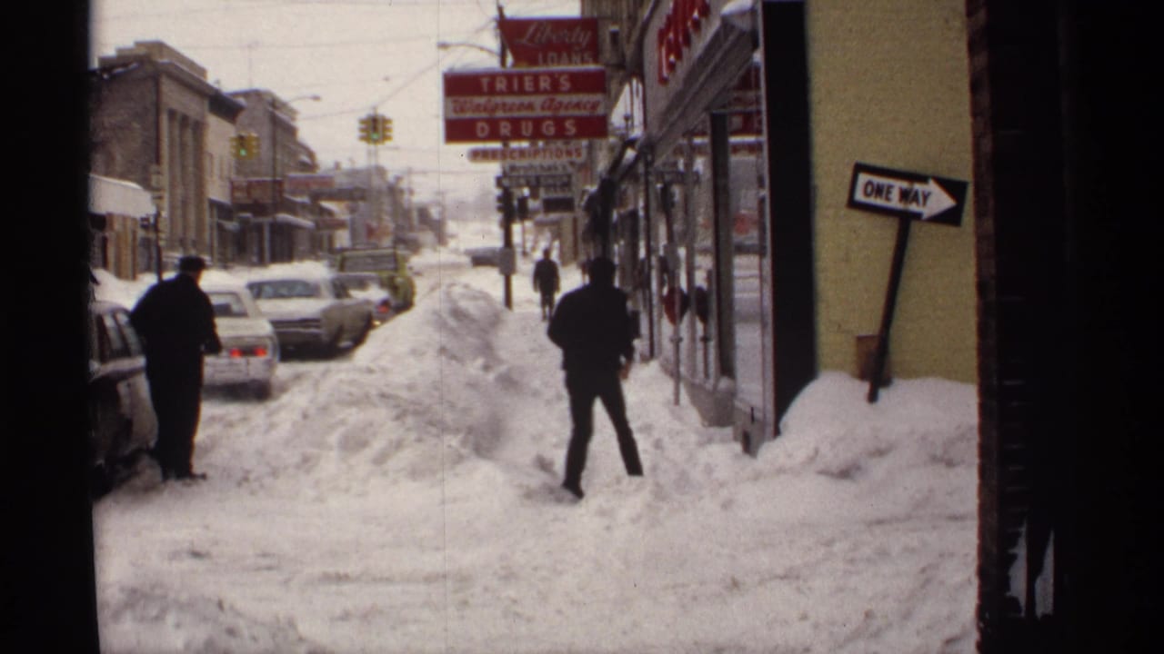 Bundled pedestrians walk near parked cars on a snowy street lined with storefronts.