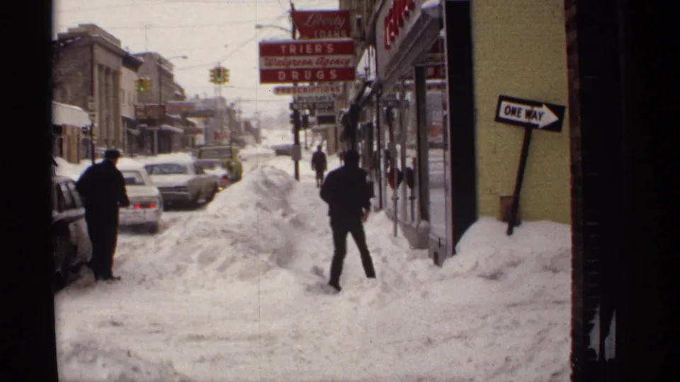 1971 Wisconsin winter street with a pedestrian crossing through snow, parked cars, storefront signs, and a leaning one-way sign.