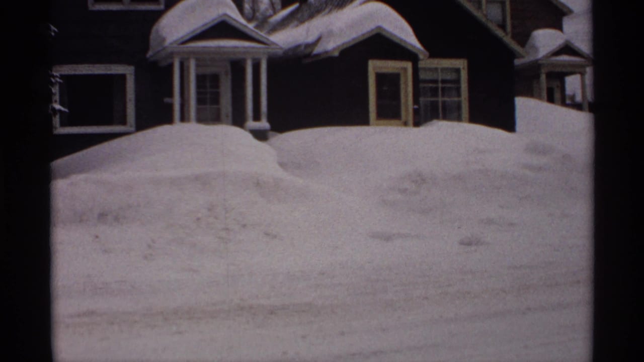 1971 Wisconsin 8mm frame showing a deep roadside snowbank partly blocking a dark residential house with white trim.