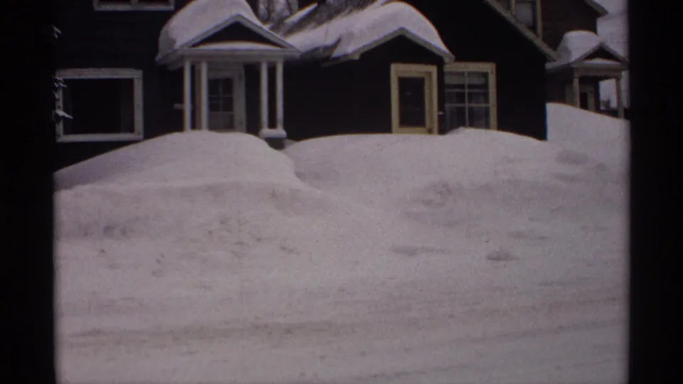1971 Wisconsin 8mm frame showing a deep roadside snowbank partly blocking a dark residential house with white trim.