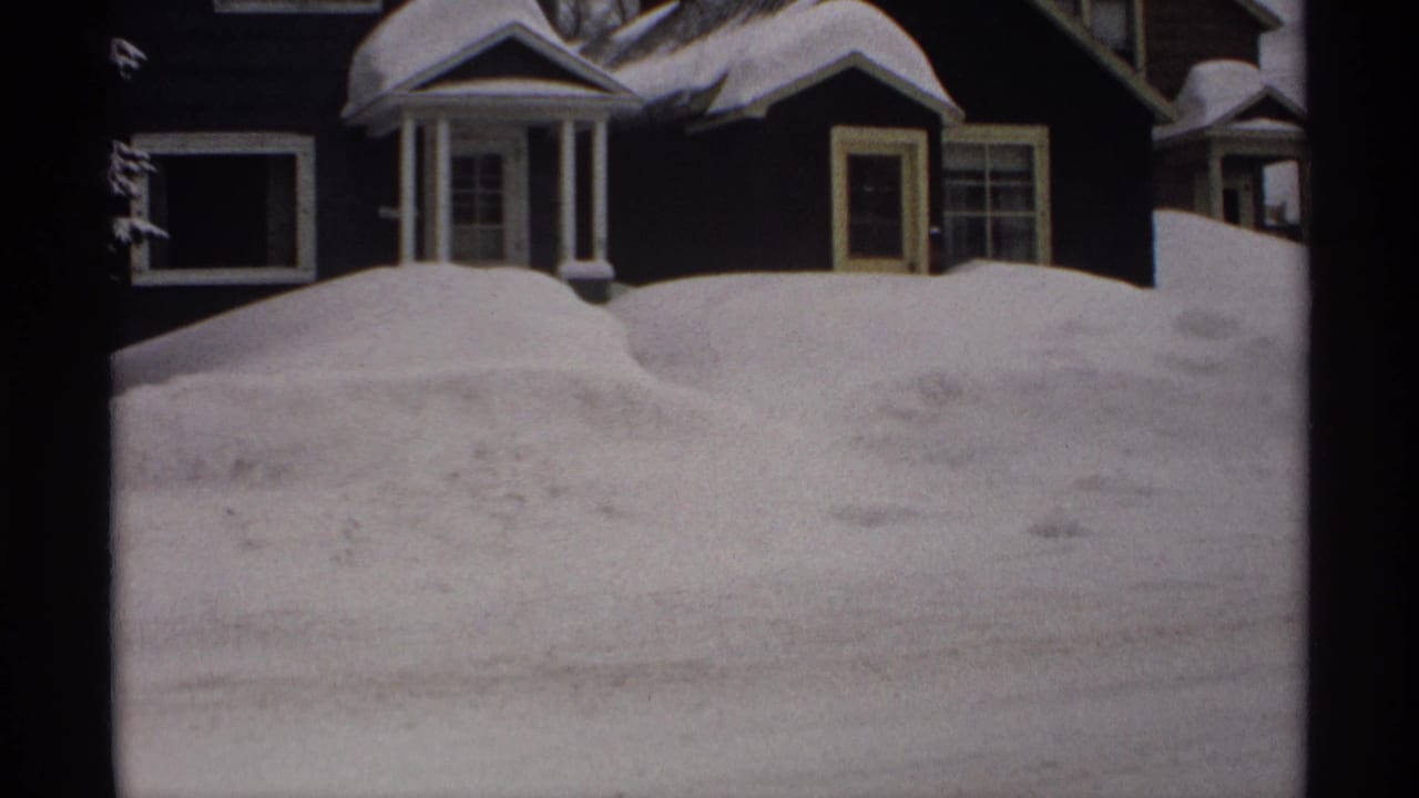 Snowbank and dark house facade with porch columns, door, window, and snowy roofline.
