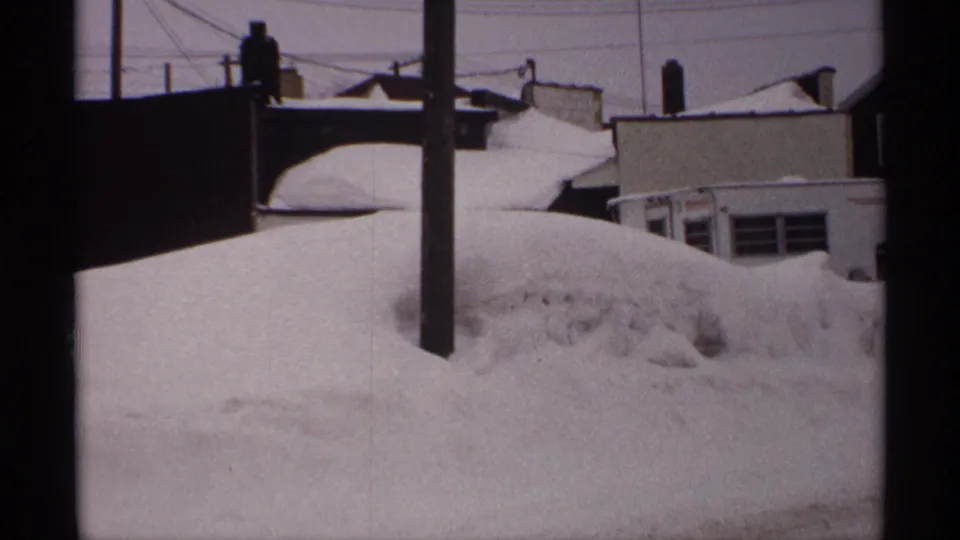 1971 Wisconsin home-movie frame looking past a utility pole toward rooftops and heavy snowdrifts along a quiet street.