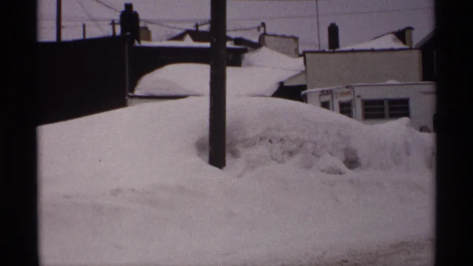 1971 Wisconsin 8mm frame of a snowbound residential street edge with a utility pole, rooftops, and piled drifts.
